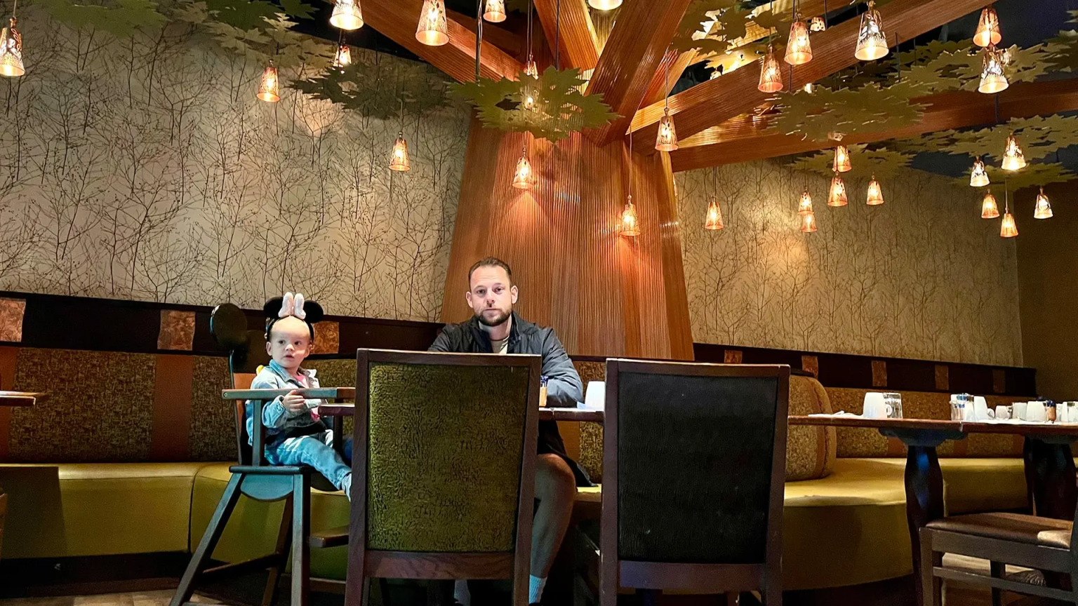 Ontbijt in de Golden Forest Club lounge van Sequoia Lodge in Disneyland Parijs, met een man en een kind in een kinderstoel aan een gedekte tafel onder een opvallende boom-vormige lichtarmatuur.
